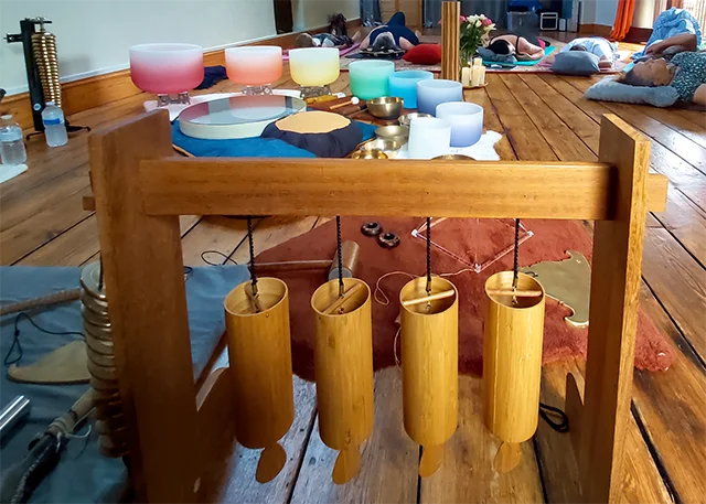 Koshi chimes hanging from a wooden stand, with singing bowls and people resting during a sound bath beyond