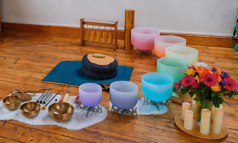 Crystal and metal singing bowls arranged on a wooden floor with a cushion, flowers and candles