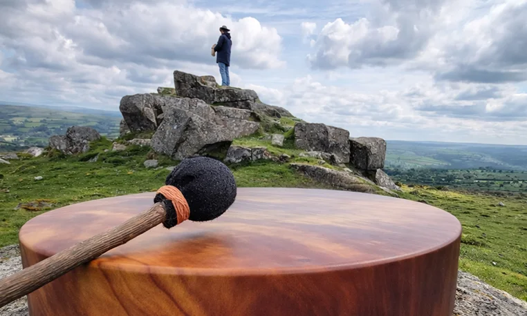 A frame drum and beater resting on a Dartmoor tor, with a figure standing on the rocks beyond