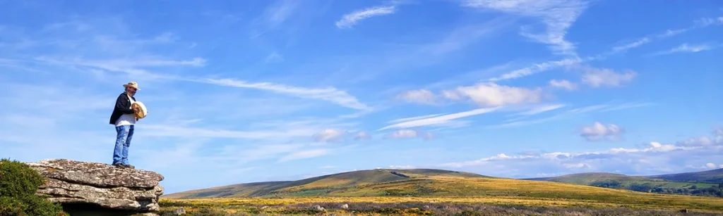 James Schindler-Ord standing on a granite tor playing a frame drum under a wide blue Dartmoor sky