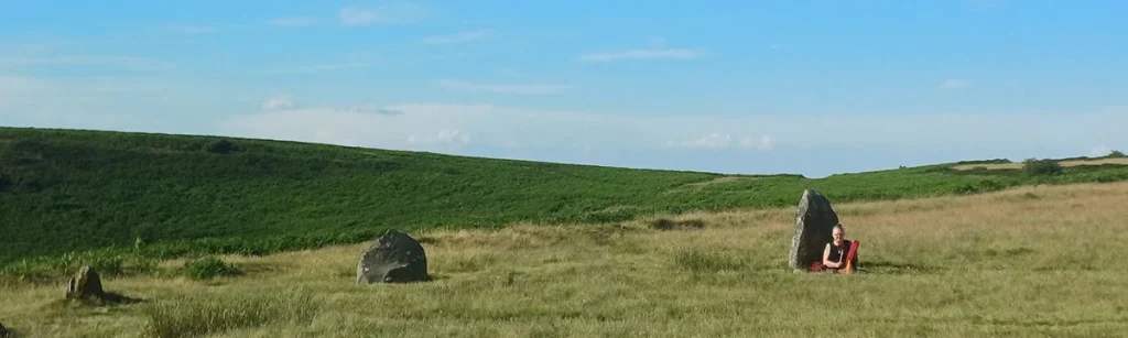A figure sitting against a standing stone in moorland grass under a blue sky